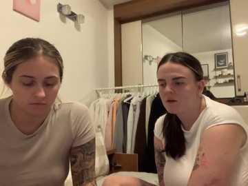 Media: Video of two young women with light skin and medium-length dark hair, wearing casual white t-shirts, sitting in a cluttered bedroom with clothes hung on racks and a wooden mirror.