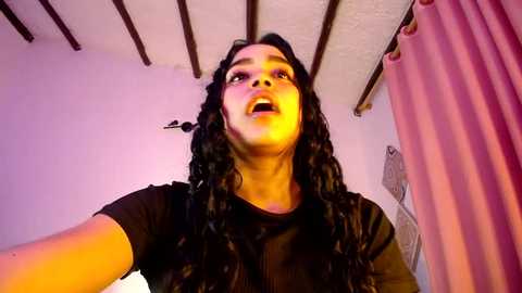 Media: Video of a woman with long, curly black hair, wearing a black top, taken from a low angle, showing her face and upper body, in a room with wooden beams and pink curtains.