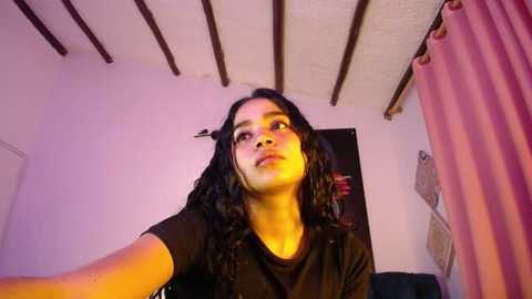 Media: Video of a young South Asian woman with medium skin tone, long black hair, and medium build, wearing a black shirt, captured from a low angle, in a room with exposed wooden beams, white walls, and pink curtains.