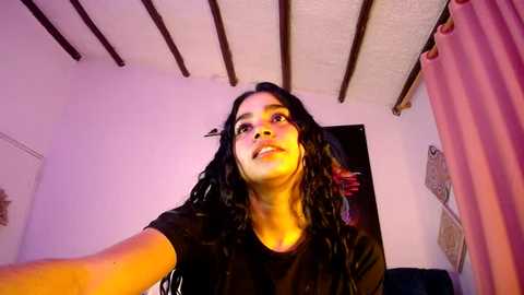 Media: Video of a young woman with medium skin tone, long black hair, wearing a black shirt, standing in a room with white walls and exposed wooden beams. She's smiling and taking a selfie.