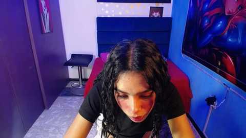 Media: Video of a young woman with long black braids, wearing a black top, leaning over a red sofa in a dimly lit room with blue walls and art on the right.