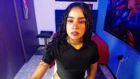 Media: Video of a young woman with long curly black hair, wearing a black ribbed crop top, standing in a brightly lit room with blue walls, a red chair, and a TV showing a colorful anime scene.