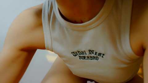 Media: Video of a woman wearing a tight, white tank top with \"World's Best Beer\" logo. Her upper body, including shoulders and collarbone, is visible, highlighting her slender physique. Background is blurred, focusing attention on the tank top.