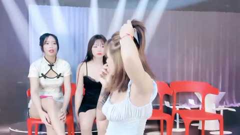 Media: Video of three Asian women in a studio; one in white dress, one in black lingerie, and one in black dress, seated on red chairs; spotlight on blonde woman in white dress.