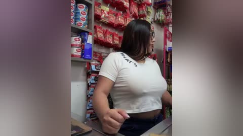 Media: A video of a Latina woman with long dark hair, wearing a white cropped top and blue jeans, working behind a checkout counter in a convenience store. The background features shelves of snacks and drinks.