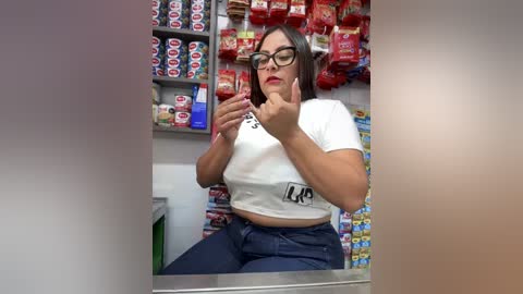 Media: Video of a Latina woman with glasses, medium build, wearing a white crop top and blue jeans, in a convenience store, pointing at a soda can.