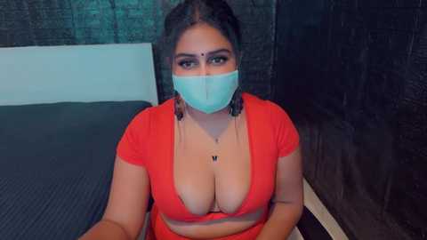 Media: Video of a South Asian woman with fair skin, large breasts, and dark hair, wearing a blue surgical mask and red crop top, sitting on a dark blue mat in a dimly lit room with dark walls.