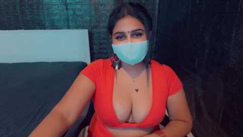 Media: Video of a South Asian woman with medium skin tone, wearing a red crop top that reveals cleavage, a blue mask, and a bindi. She sits on a bed with dark bedding in a dimly lit room.