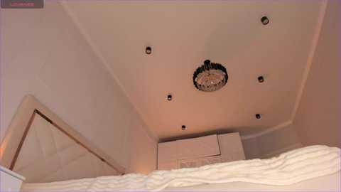 Media: Video of a modern, minimalist bedroom ceiling with five recessed lights, a chandelier, and a white wall. A white bed with a white quilted headboard is partially visible in the lower left corner.