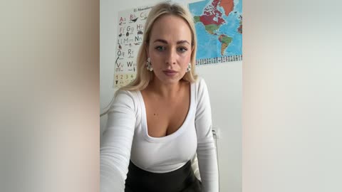 Media: Video of a blonde woman with fair skin, wearing a white long-sleeve top and black skirt, sitting against a white wall adorned with educational posters.