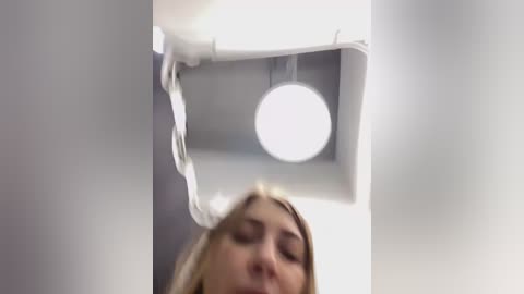 Media: A video showing a young blonde woman with closed eyes, positioned below a white, rectangular ceiling fixture with a circular light bulb. The image has a soft focus and is taken from a low angle.