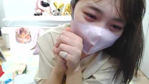 Video of an East Asian woman with long black hair, wearing a light pink face mask, holding her hands together in a white room cluttered with stuffed toys and a window.