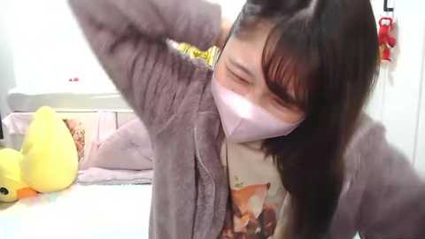 Media: Video of an Asian woman with long black hair, wearing a gray cardigan, white mask, and yellow T-shirt, stretching her arm while leaning against a bed with a yellow stuffed animal. Background includes a white wall and soft furnishings.