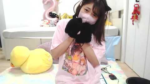 Video of a young woman with dark hair and a light pink t-shirt featuring anime characters, wearing a face mask, kneeling in a brightly lit room with a white bathtub and a plush yellow duck.