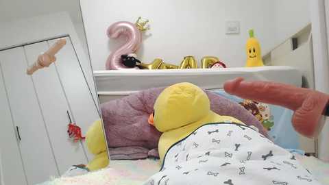 Media: Video of a baby in a yellow onesie with animal prints, lying on a bed with a plush toy and a mirror, surrounded by colorful toys and a large, erect penis.