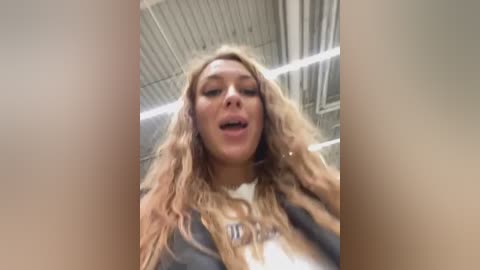 Media: A video of a blonde woman with wavy hair, wearing a gray shirt, captured from a low angle, showing her smiling face and part of the industrial ceiling.