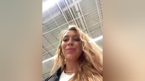 Media: Video of a young woman with long, wavy blonde hair, wearing a black jacket and a silver necklace, standing indoors with a high ceiling and visible ductwork.