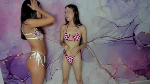 Video of two young women in matching red and white heart-print bikinis, one adjusting her bikini top, against a mural backdrop with purple and pink tones.