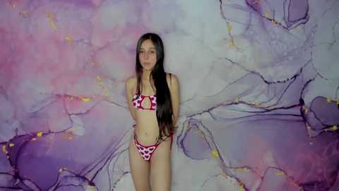 A video of a young, slim woman with long brown hair in a red and white polka-dot bikini, posing against a swirling, pastel-hued, abstract background.