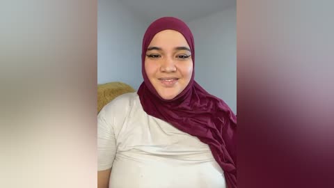 Video of a smiling, light-skinned woman with a round face, wearing a white t-shirt and a maroon hijab, seated indoors against a plain, light-colored background.