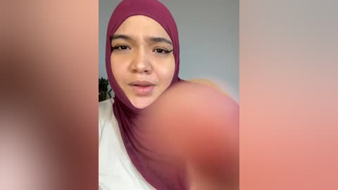 Video of a woman with medium skin tone, wearing a maroon hijab and white top, blurred background, focusing on her face.