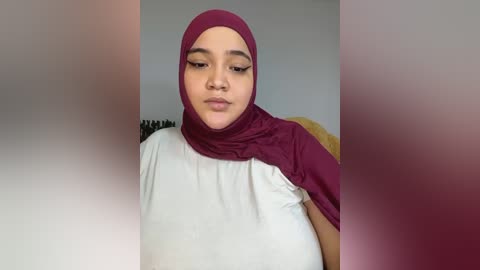 Video of a young woman with light brown skin, wearing a white shirt and maroon hijab, sitting indoors with blurred background.