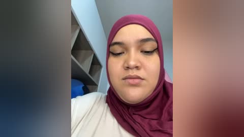 Video of a young woman with light brown skin, wearing a maroon hijab and white top, looking down with neutral expression. Background features a blue wall and open shelves.