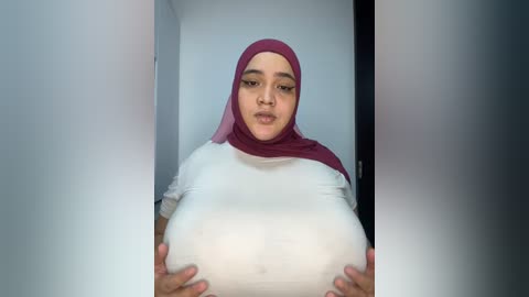 Media: Video of a pregnant woman with medium skin tone, wearing a white t-shirt and a pink hijab, holding her large, round belly against her chest.