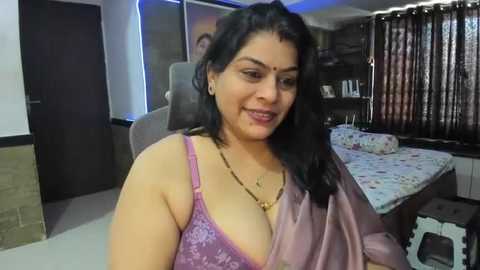 Media: Video of a South Asian woman with medium skin tone, black hair, and wearing a pink lace bra, smiling in a modern bedroom with a bed, curtains, and wooden furniture.