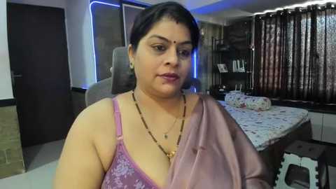 Media: Video of a middle-aged South Asian woman with medium skin tone, wearing a pink floral bra and a draped purple saree, sitting in a modern living room with dark wooden furniture and a floral tablecloth.