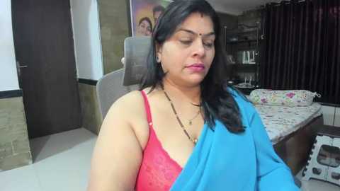 Media: Video of a South Asian woman in a red bra, blue sari, and gold necklace, sitting in a modern bedroom with a brown bed and floral sheets, a dark curtain, and a TV in the background.