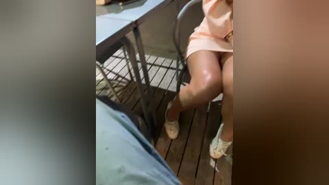 Media: Video of a woman in a peach dress sitting on a patio with a glass table and metal chairs. Her legs are crossed, and she wears beige shoes.