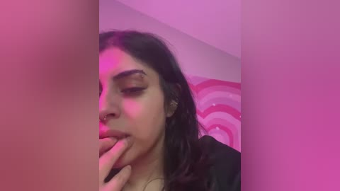 Media: Video of a young Latina woman with long dark hair, wearing a black top, sensually biting her finger under pink and purple lighting. Background features a pink, striped wall.