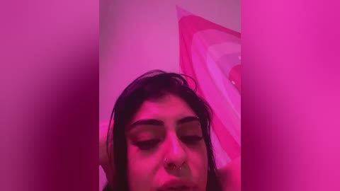 Media: Video of a woman with medium skin tone and dark hair, wearing a nose ring, eyes closed, in a room lit with pink lighting, creating a dreamy, ethereal atmosphere.