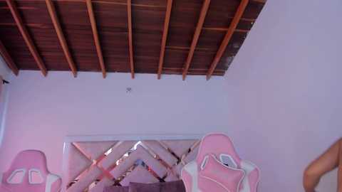 Media: Video of a bedroom with a high ceiling, wooden beams, white walls, and a diamond-patterned headboard. Pink and white pillows are visible on the bed.