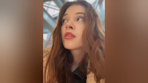 Video of a young woman with long, wavy brown hair, fair skin, and full lips, wearing a beige puffer jacket, gazing upwards in a thoughtful manner.