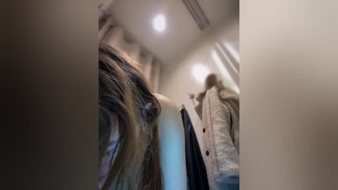 Video of a blurred, slightly tilted perspective showing a person's shoulder and back in a hallway with beige walls, a white coat, and a dark jacket hanging on a hook.