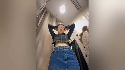 Video of a young woman with fair skin, medium build, and light brown hair, wearing a cropped dark sweater and high-waisted blue jeans, posing confidently in a dressing room with beige walls and a white coat hanging nearby.