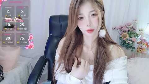 Media: Video of an East Asian woman with long brown hair, wearing off-shoulder white top and large white earrings, sitting on a black chair in a room with a floral arrangement, overlaying a digital game screen.