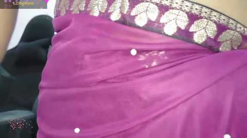 Media: A close-up video of a woman's midsection in a bright pink sari with golden floral embroidery and small white sequins, partially draped over a black chair.