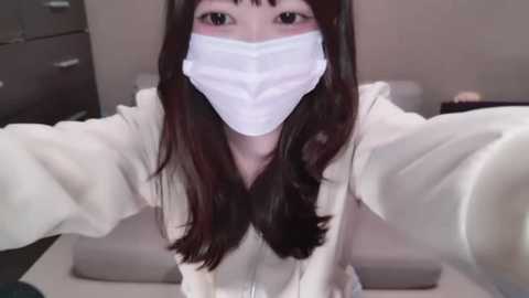 Media: A video of an Asian woman with long, dark hair, wearing a white face mask, white hoodie, and holding a camera. Background shows a beige room with a dresser.