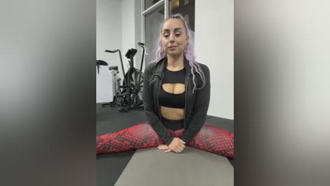 Media: Video of a woman with light skin and lavender hair, sitting in a yoga pose on a gym mat, wearing a black sports bra and red patterned leggings. Background features gym equipment and white walls.