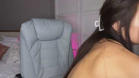 Media: A video shows a woman with long, dark hair, wearing a white headband, lying on a light blue, quilted chair in a dimly lit bedroom with soft pink lighting.