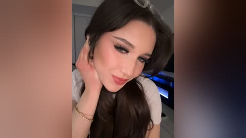 Media: Video of a young woman with long, dark hair, wearing makeup, smiling, and touching her face, blurred background.