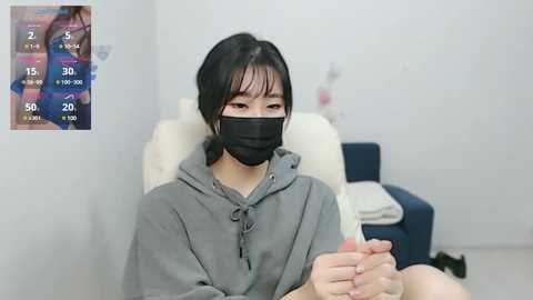 Media: Video of an Asian woman with black hair, wearing a grey hoodie and black mask, sitting in a medical chair, surrounded by charts.