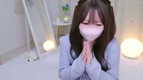 Media: Video of an Asian woman with long black hair, wearing a pink mask and a light blue sweater, kneeling with hands clasped in prayer, in a minimalist, white room with a small table and glowing spherical lamps.