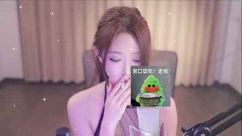 Media: Video of a fair-skinned, slim Asian woman with long brown hair, wearing a white sleeveless top, sitting at a microphone in a beige room with curtains. She has her eyes closed and is covering her mouth with her hands. A green frog emoji is superimposed on her face.