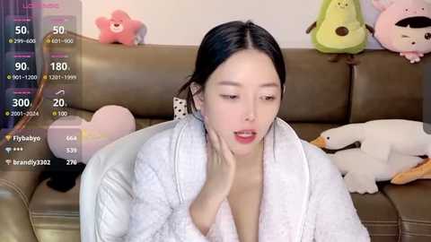 Media: Video of an Asian woman in a white bathrobe, sitting on a brown couch. Background features plush toys, including a pink bear, a green frog, and a white dog.