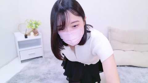 Media: Video of a young Asian woman with light skin, straight black hair, and a white face mask, kneeling on a grey rug in a minimalist room with a beige sofa and white shelves.