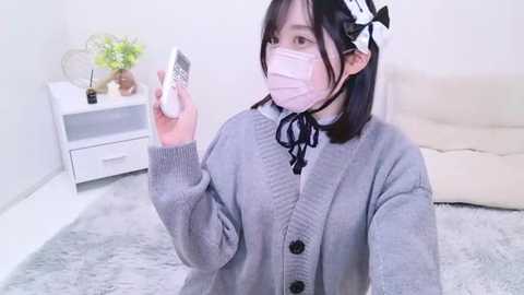 Media: Video of an Asian woman with pale skin, wearing a light grey cardigan, black bow tie, and pink mask, holding a phone, in a modern, minimalist room with white walls and a beige couch.